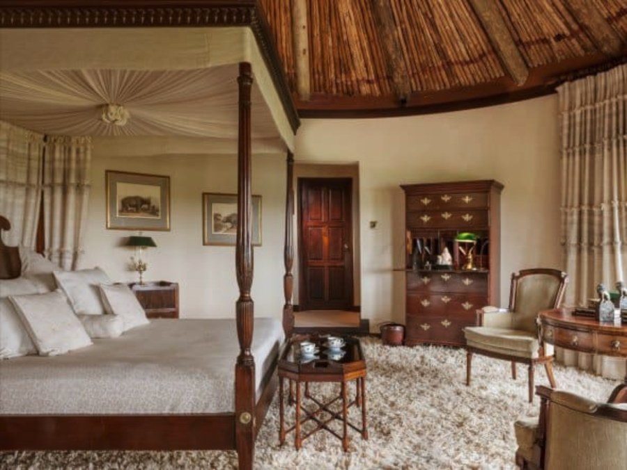 Bedroom at Ol Jogi with a four-poster bed, wooden cabinets, armchairs and a shaggy carpet under a thatched roof