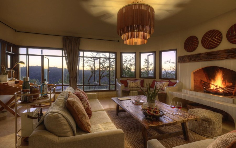 Sitting room at Ol Lentille with sofas, a lit fireplace and a curve of windows looking out over hills at dusk