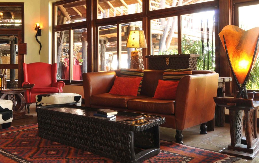Lounge at Ol Tukai Lodge with a leather sofa, a red wingback chair, a carved wooden table and a patterned rug