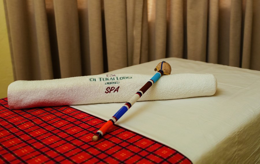 Rolled spa towel embroidered with the Ol Tukai Lodge name and a beaded stick on a massage bed with a red cloth