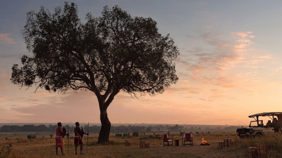 Two Maasai men by a lone tree at sunrise, beside a campfire, chairs and a safari vehicle on the Mara plains