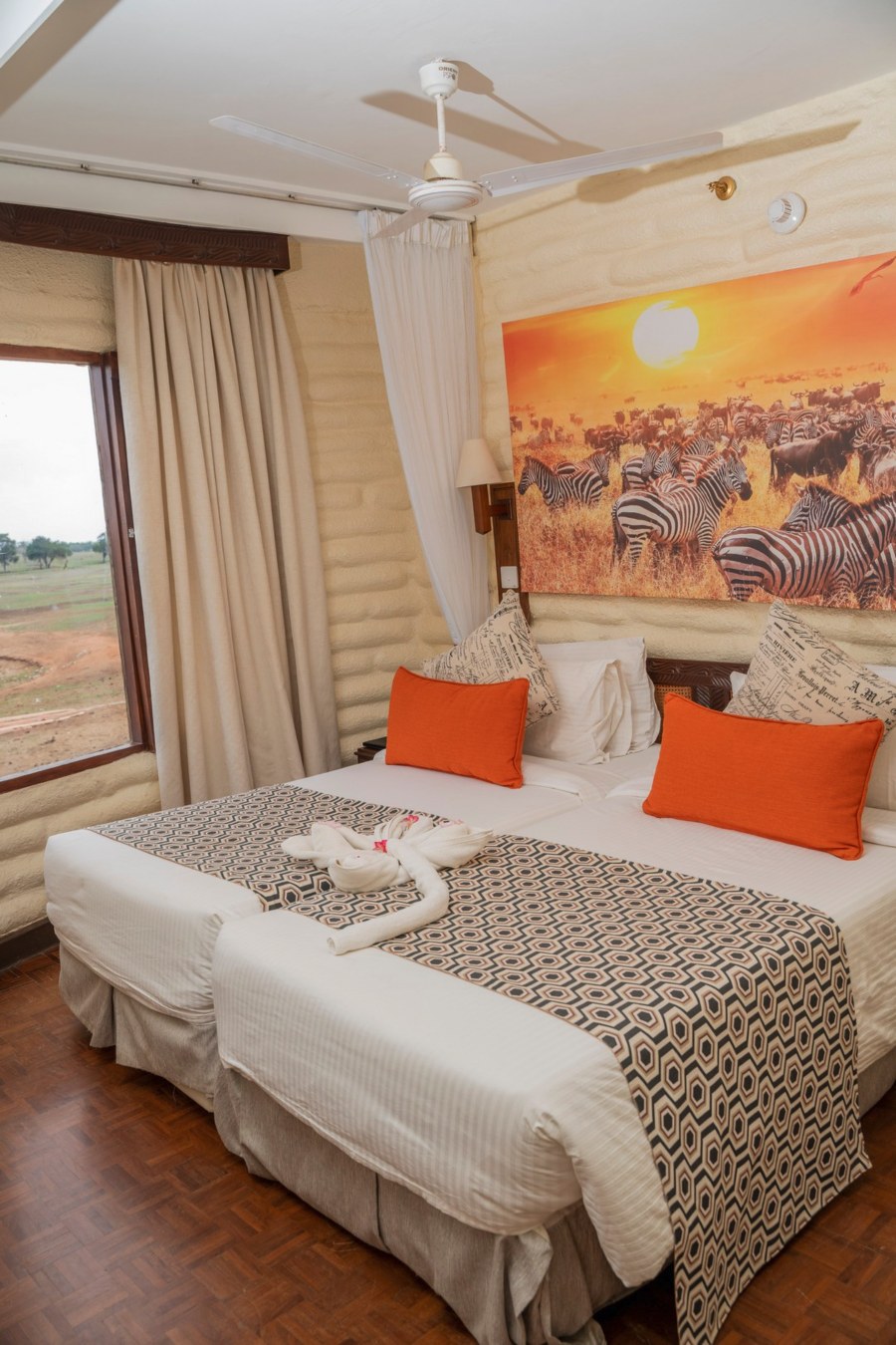 Twin beds with orange cushions under a zebra painting at Salt Lick Safari Lodge, with a window onto the plains