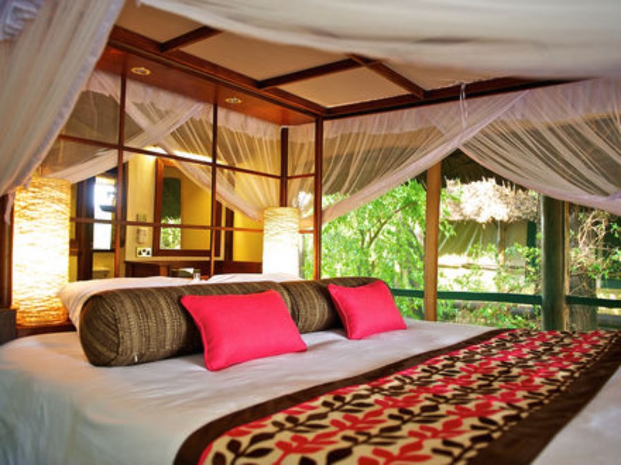Bed with a mosquito net and pink cushions in a tent opening onto trees at Samburu Intrepids