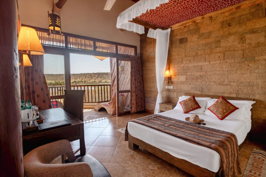 Stone-walled bedroom with a canopy bed, desk and balcony looking over bush at Samburu Simba Lodge
