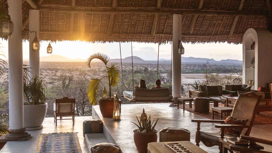 Two guests on a hanging daybed in an open thatched lounge looking over bush to hills at sunset at Sasaab