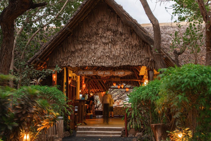 Thatched entrance with a carved Severin Safari Camp sign, lit by lanterns among trees