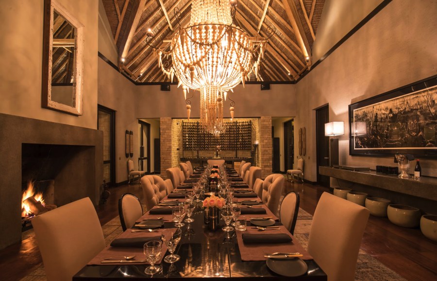 Long dining table under a beaded chandelier and thatched ceiling, beside a lit fireplace at Sirai House