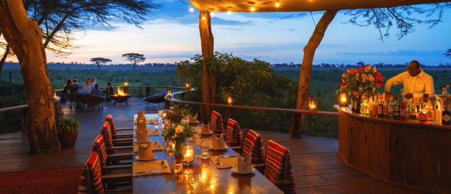 Candlelit dining table and bar on a wooden deck, with guests around a fire pit at dusk at Sirikoi