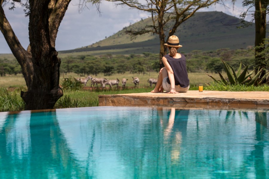 Woman sitting at the edge of a pool watching zebras graze below a hill at Sirikoi