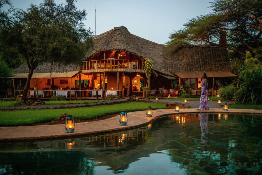 Woman standing by a lantern-lit pond in front of the thatched main lodge at dusk at Tawi Lodge