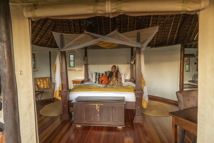 Woman sitting on a four-poster bed in a thatched round cottage with a wooden chest at Tawi Lodge