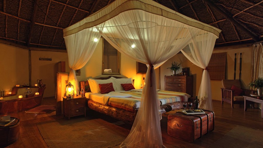 Four-poster bed with a mosquito net under a thatched roof, lit by lanterns, at Elewana Tortilis Camp Amboseli