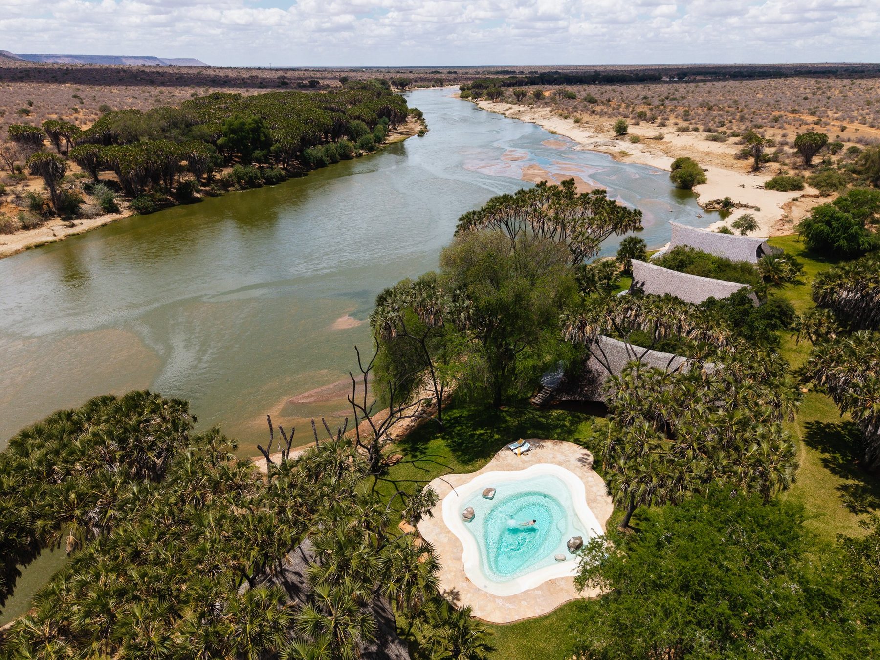 Aerial view of Galdessa Camp’s pool and roofs under green trees on the bank of the wide Galana River