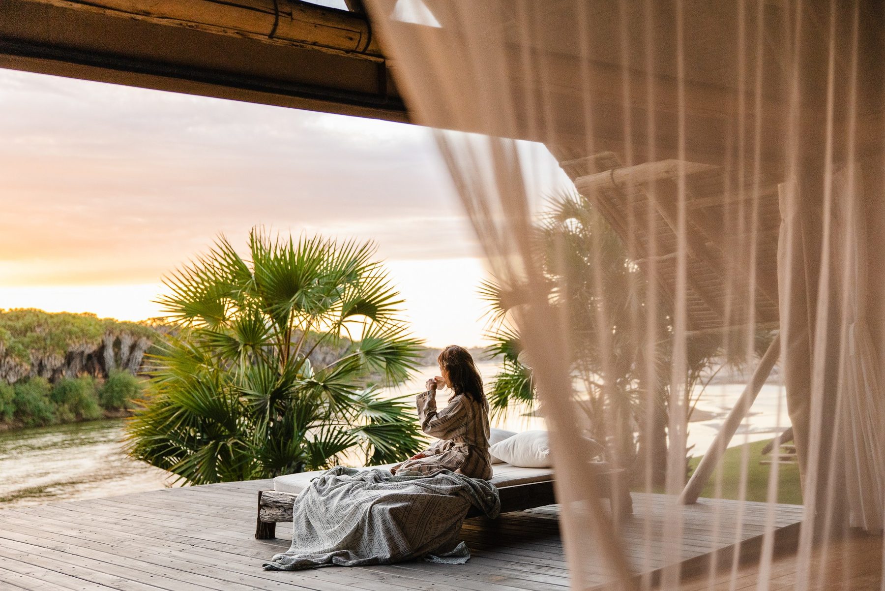 Guest sitting on a bed behind sheer curtains at Galdessa Camp, looking past palms to the river at sunset