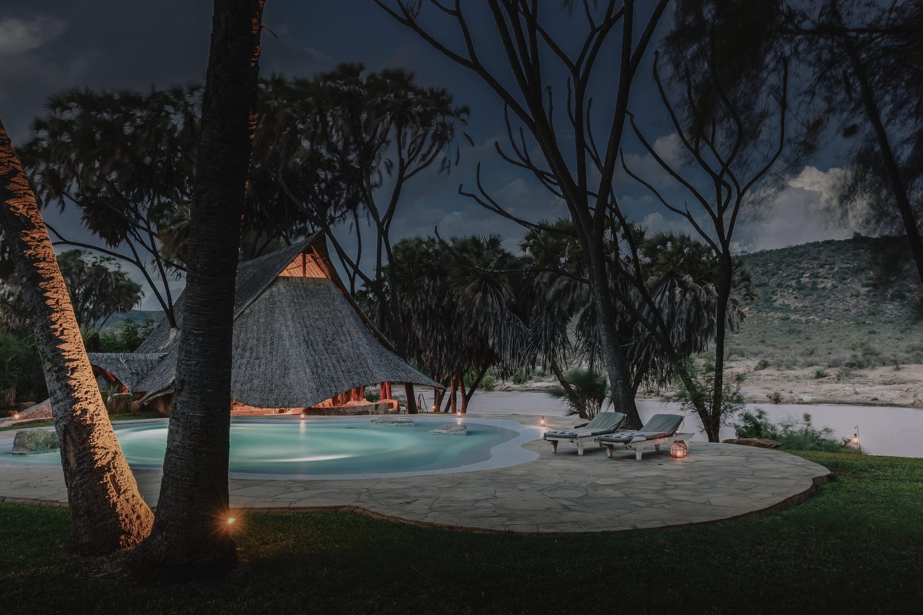 Pool and loungers lit at dusk beneath a tall thatched roof and palms at Galdessa Camp