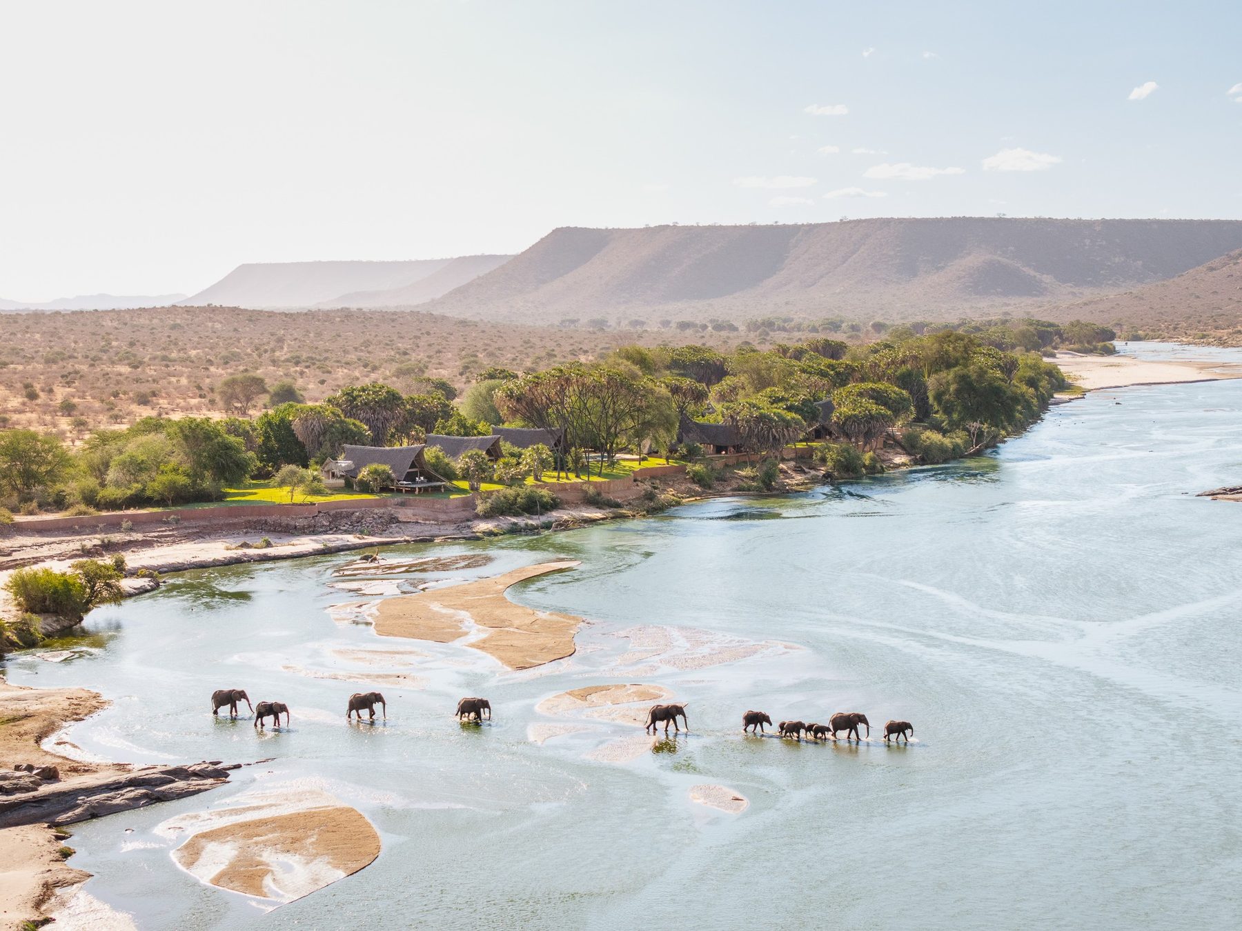Line of elephants crossing the shallow Galana River below green riverbank trees and a flat-topped ridge