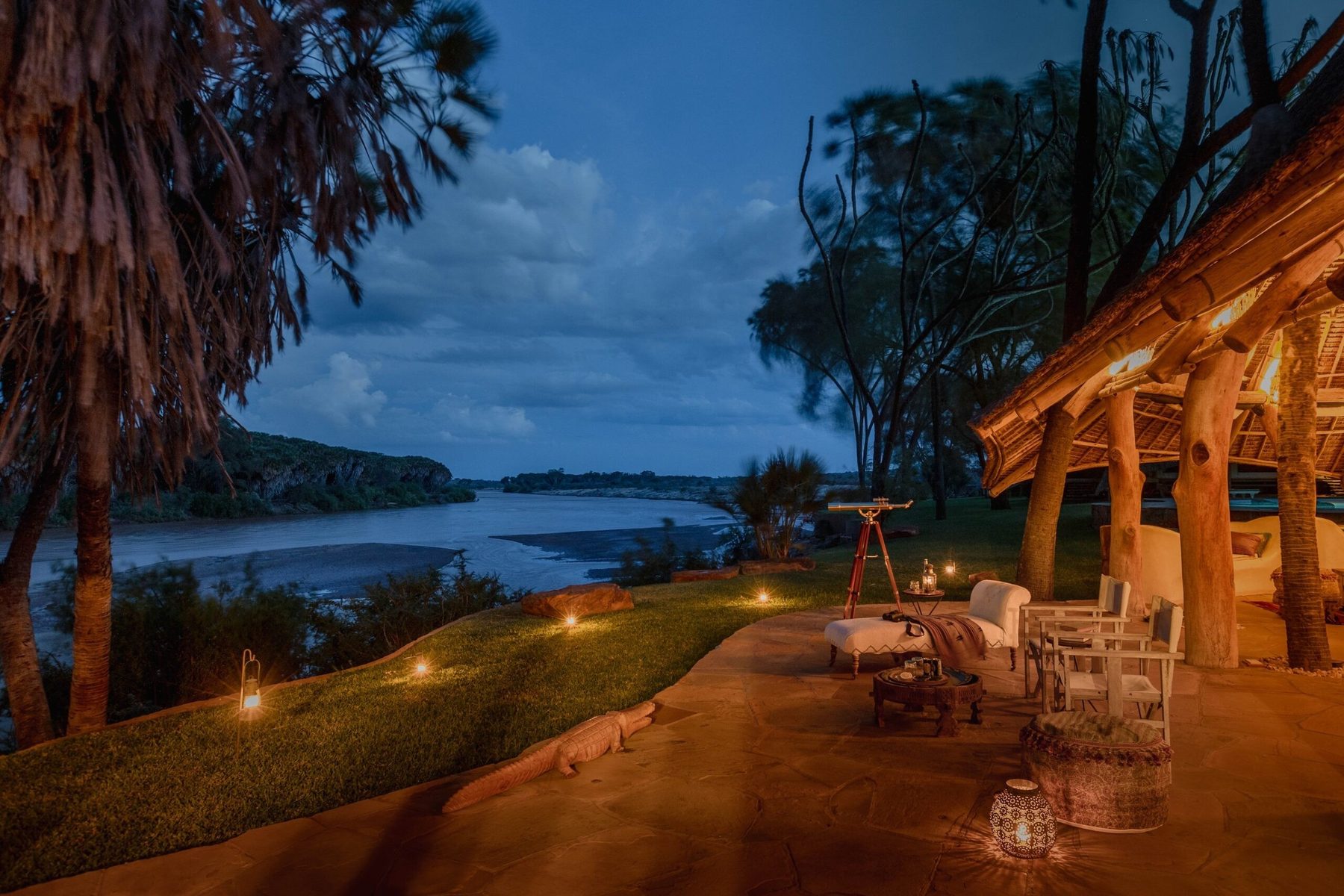Lantern-lit lawn and wooden deck with chairs above the Galana River at dusk at Galdessa Camp