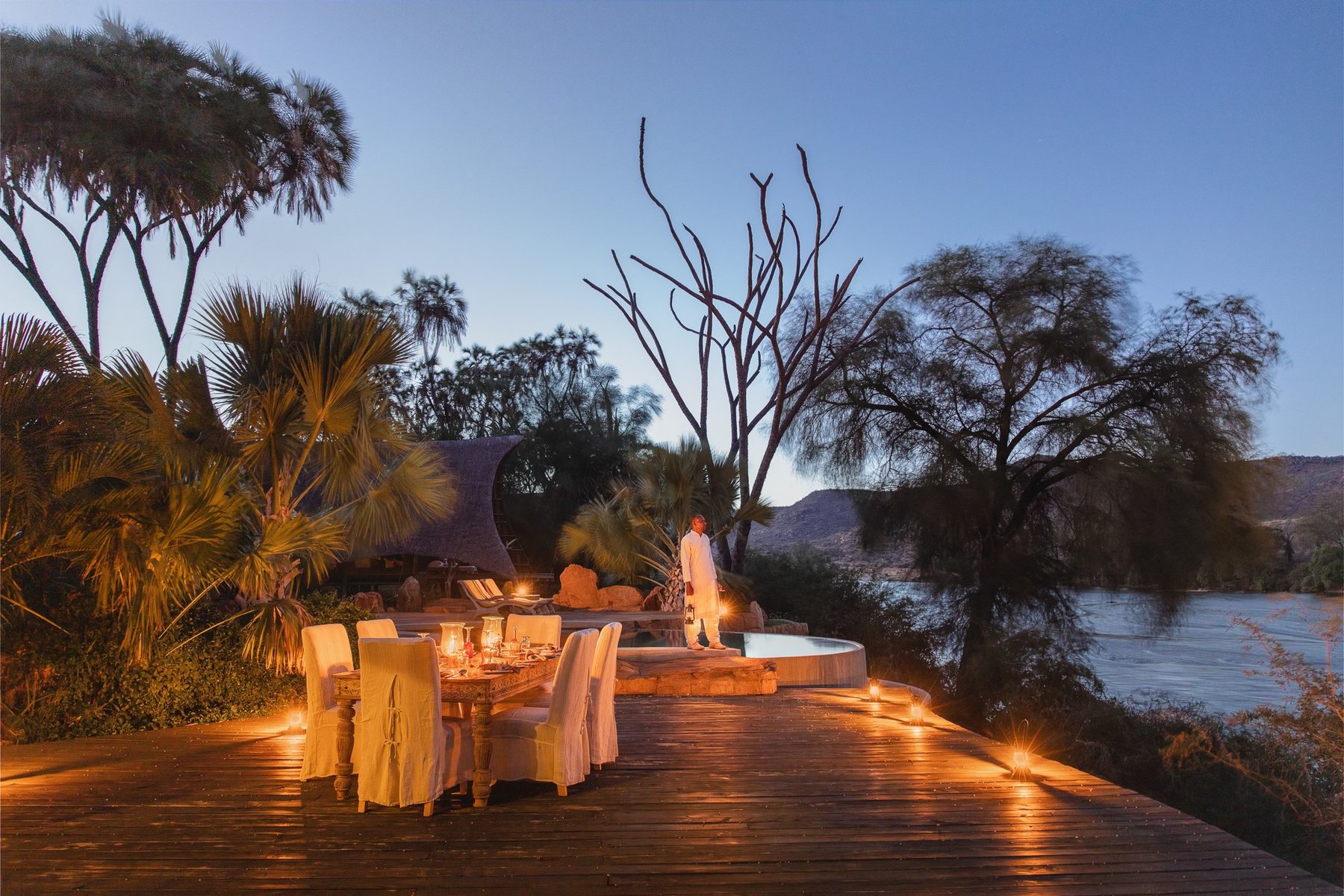 Candlelit dinner table on a deck lined with lanterns beside the river at dusk at Galdessa Camp