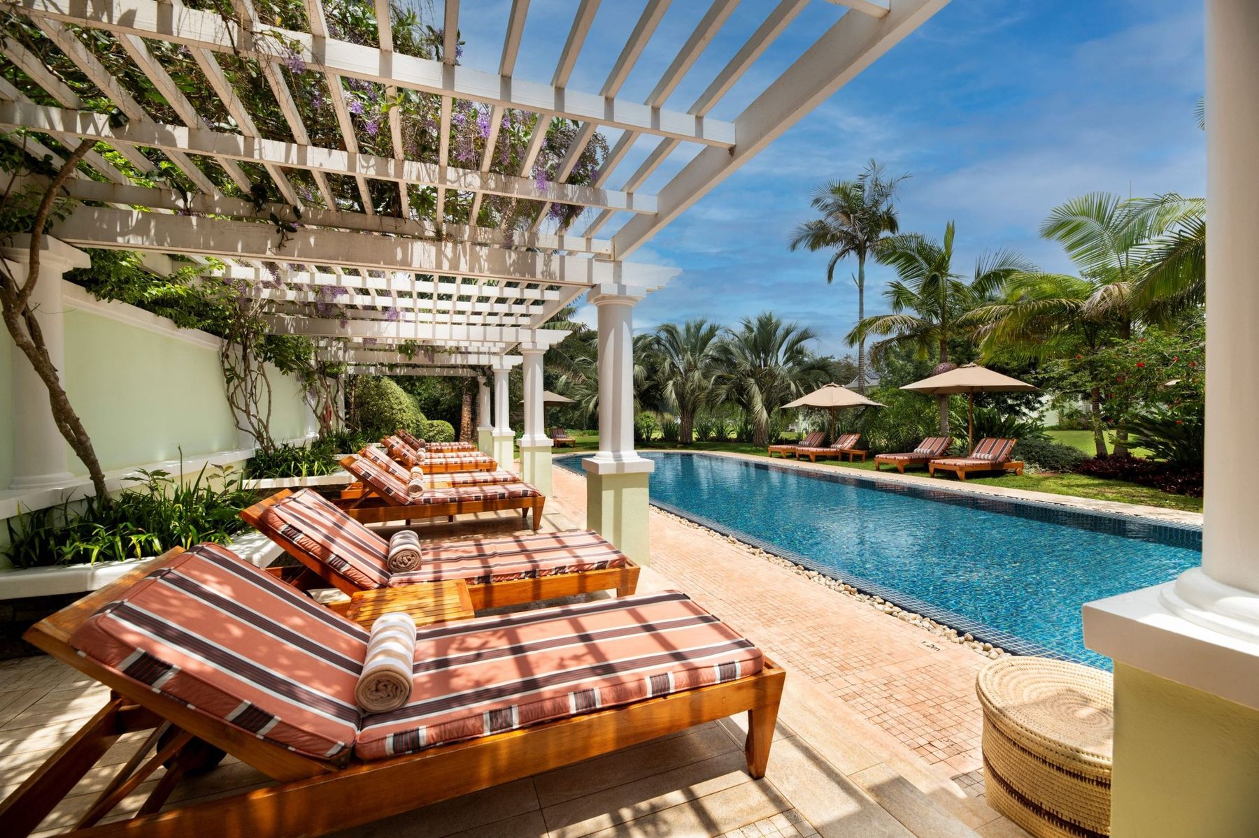 Striped sun loungers under a pergola beside the long pool at Hemingways Nairobi, with palms and lawns beyond