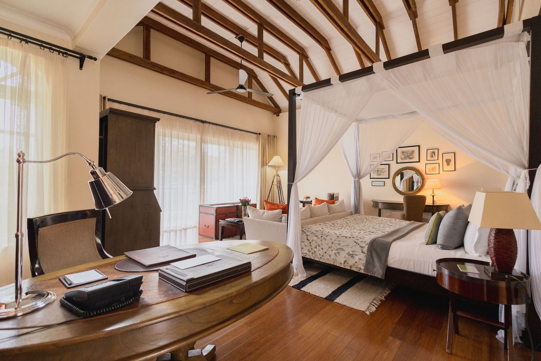 Writing desk and lamp in a Hemingways Nairobi suite, with a four-poster bed behind under a vaulted ceiling