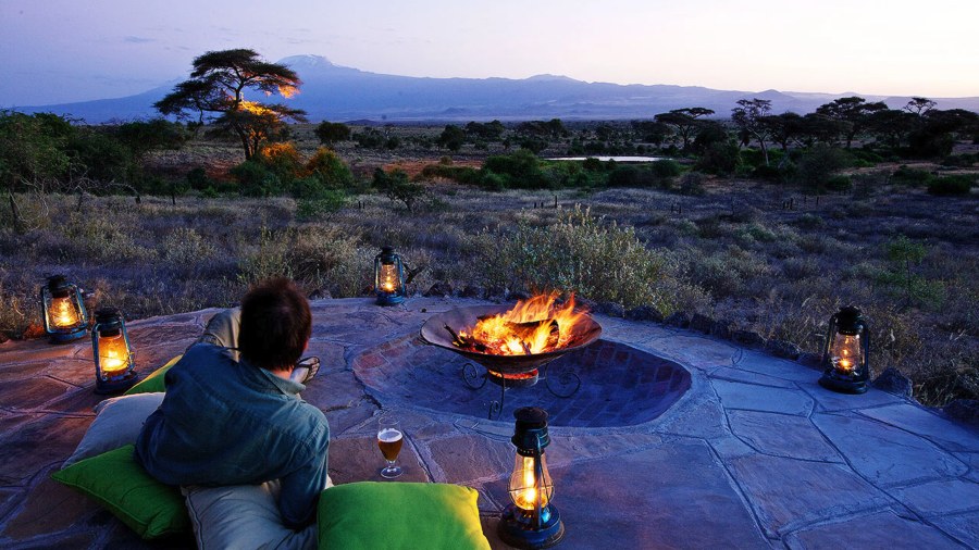 Person by a fire pit ringed with lanterns, looking over bush towards Kilimanjaro, in the Amboseli safari region