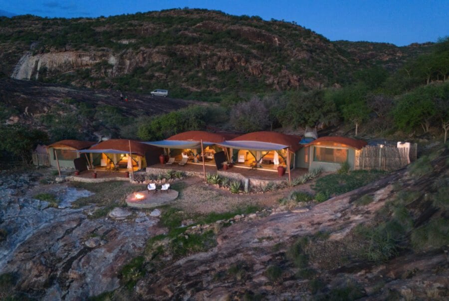 Tents with curved red roofs and a fire pit on a rocky hillside at dusk in the Kalama Conservancy