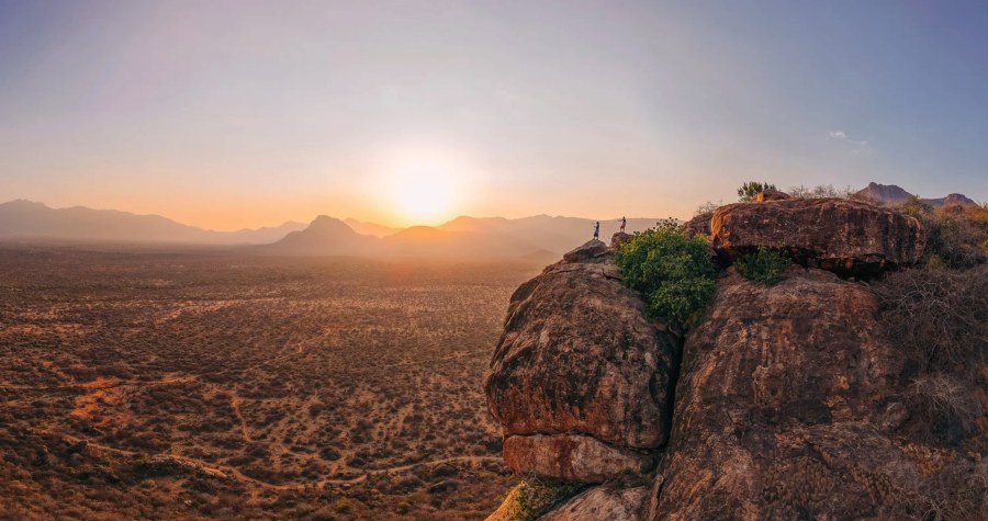 Two people on a rock outcrop above dry bush and mountains at sunset in the Namunyak Conservancy