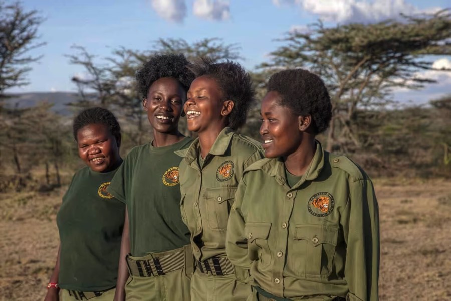 Four smiling women rangers in green uniforms standing among acacia trees in the Olderkesi Conservancy