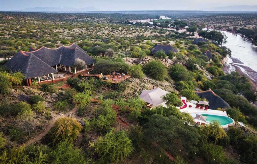Aerial view of a thatched lodge and pool on a bushy hillside above a river in the Samburu safari region
