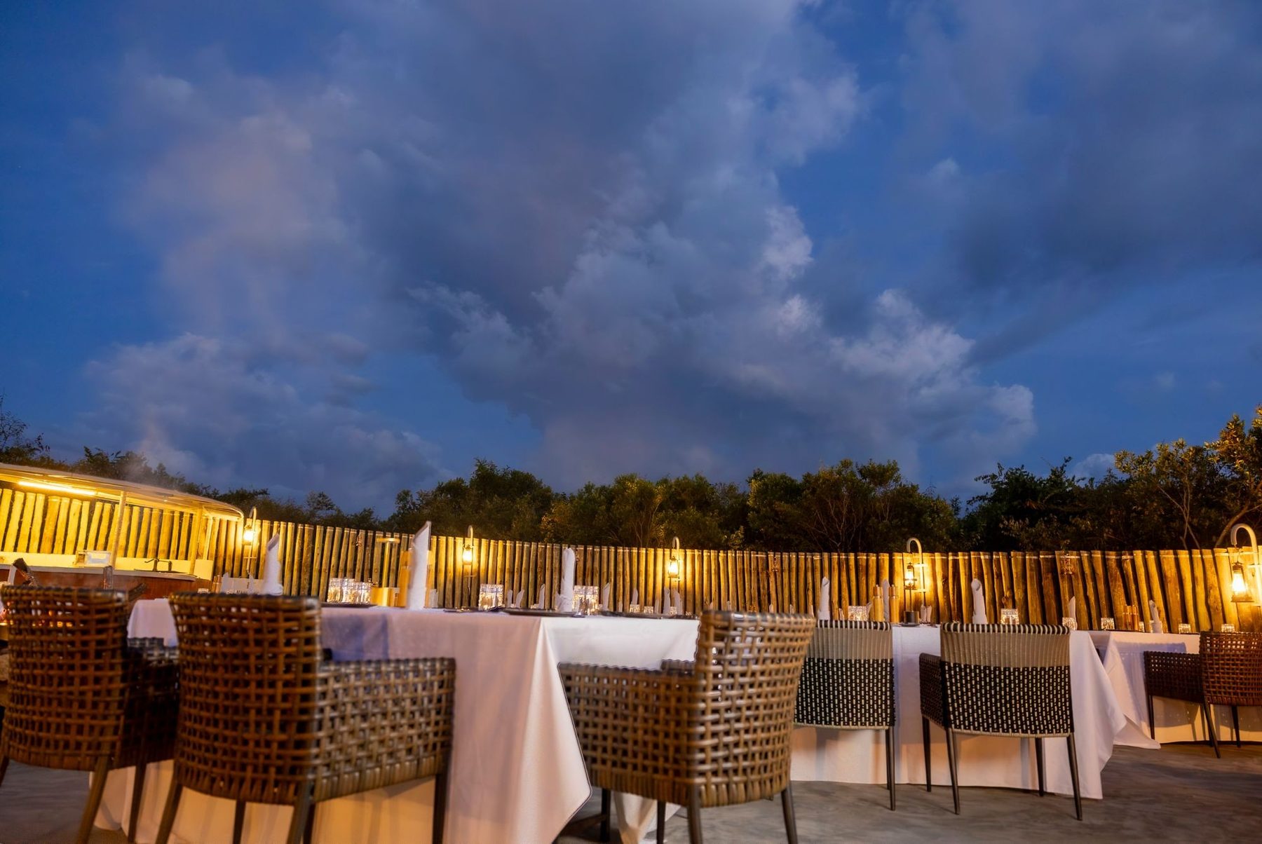 Tables laid with white linen and lantern-lit reed walls under a blue evening sky at JW Marriott Masai Mara Lodge