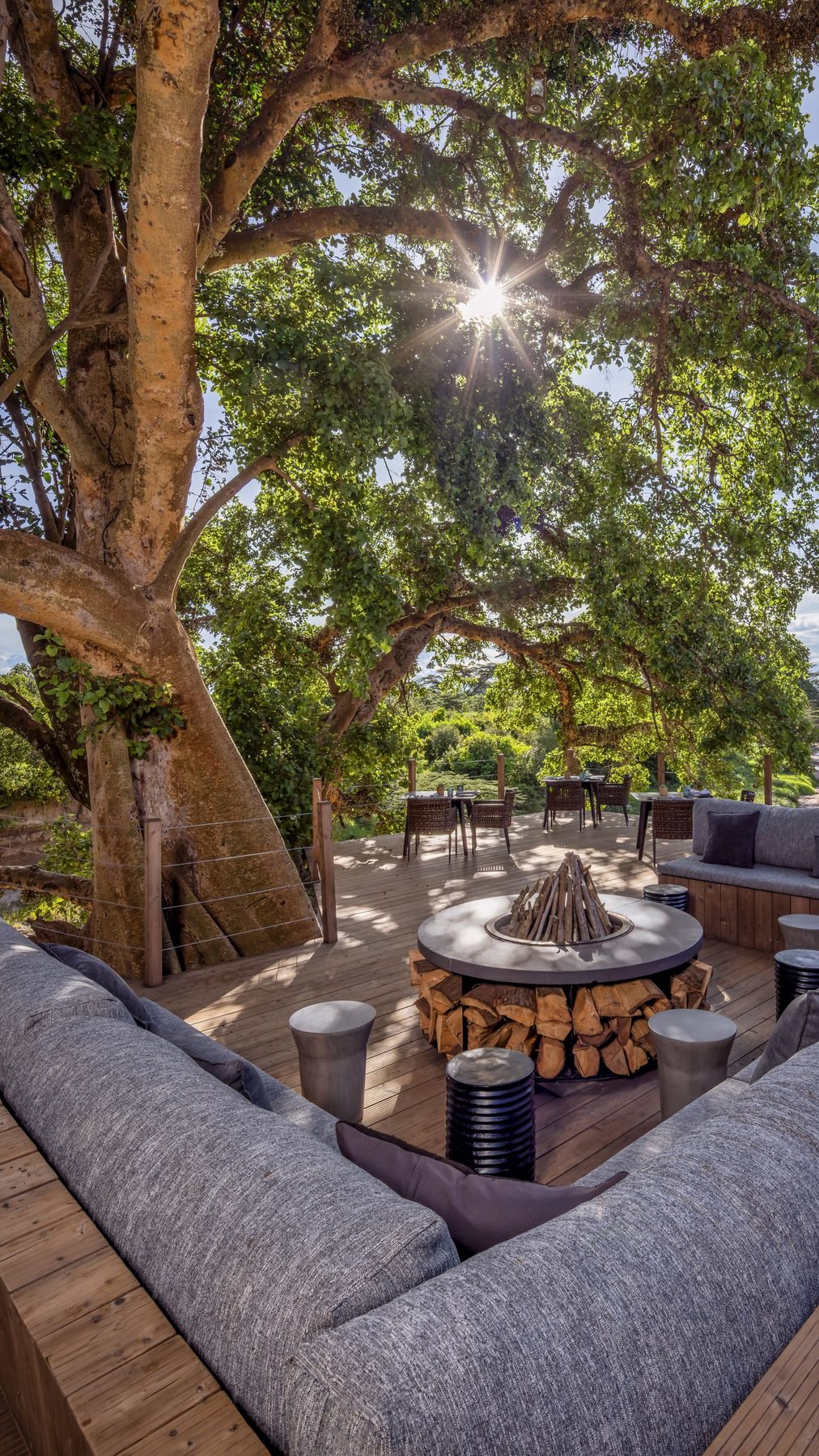 Grey sofas around a stone fire pit with stacked logs on a deck beneath a huge tree at JW Marriott Masai Mara Lodge