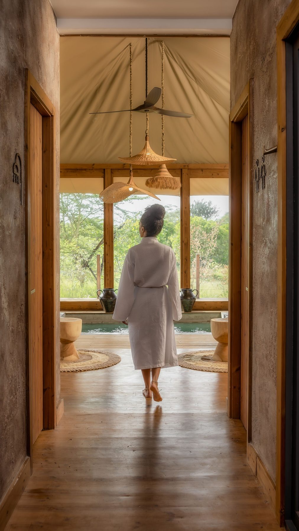 Guest in a bathrobe walking down a wooden corridor towards a suite’s bath and windows onto trees at JW Marriott Masai Mara Lodge