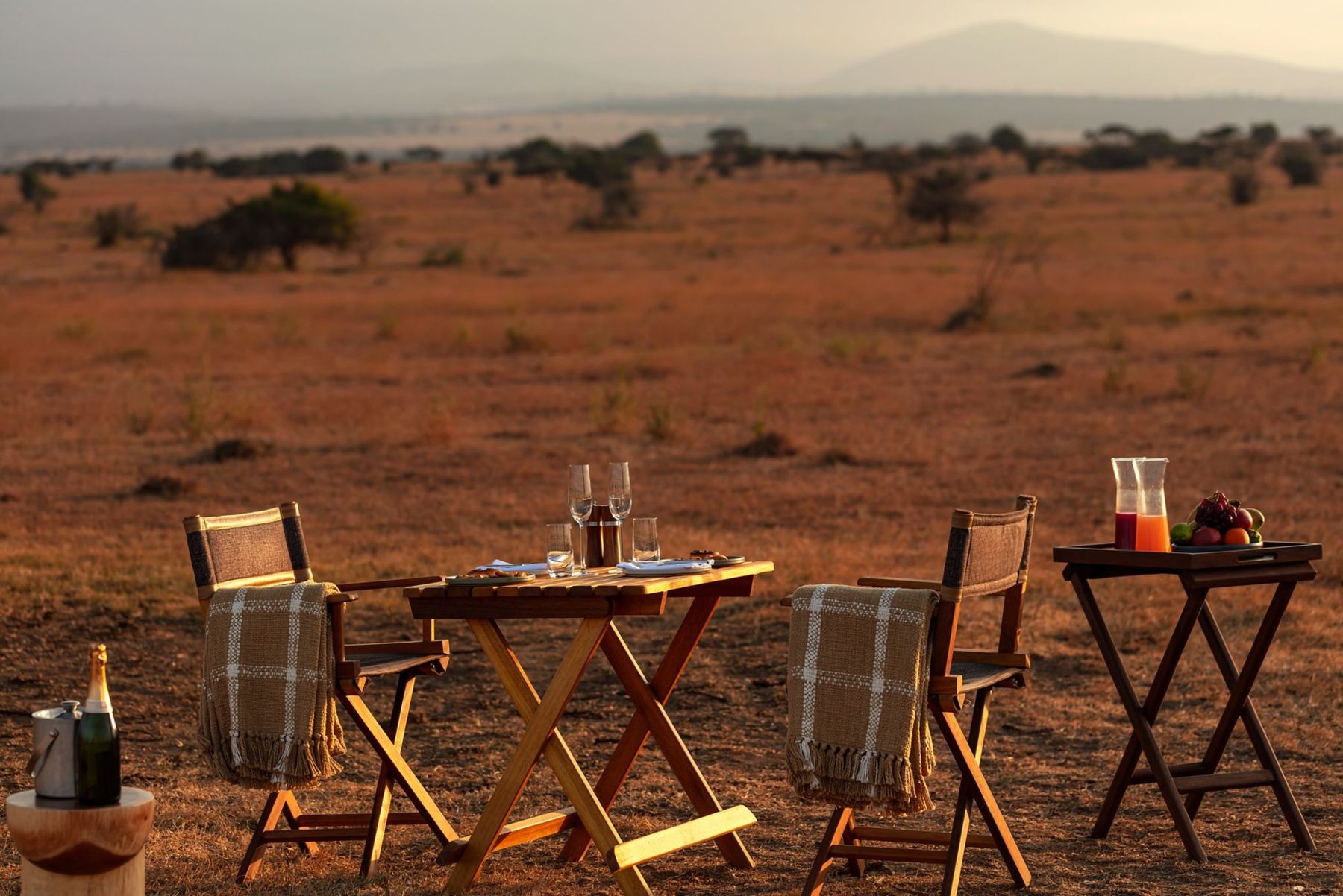 Table and safari chairs set with drinks on open red-earth plains for a bush meal from JW Marriott Mount Kenya Rhino Reserve Safari Camp