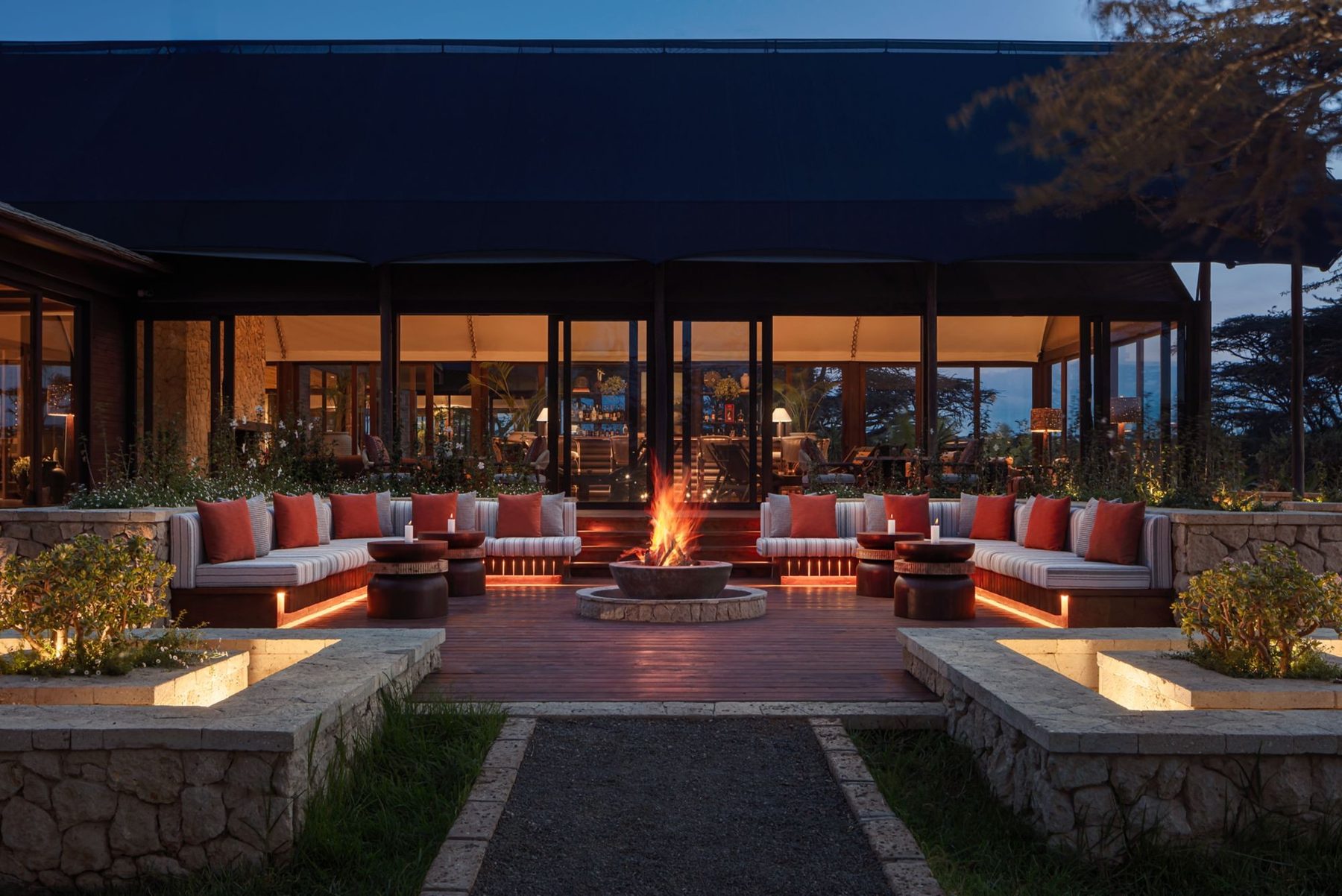 Fire pit with orange cushioned seating in front of the lit main building at JW Marriott Mount Kenya Rhino Reserve Safari Camp at dusk