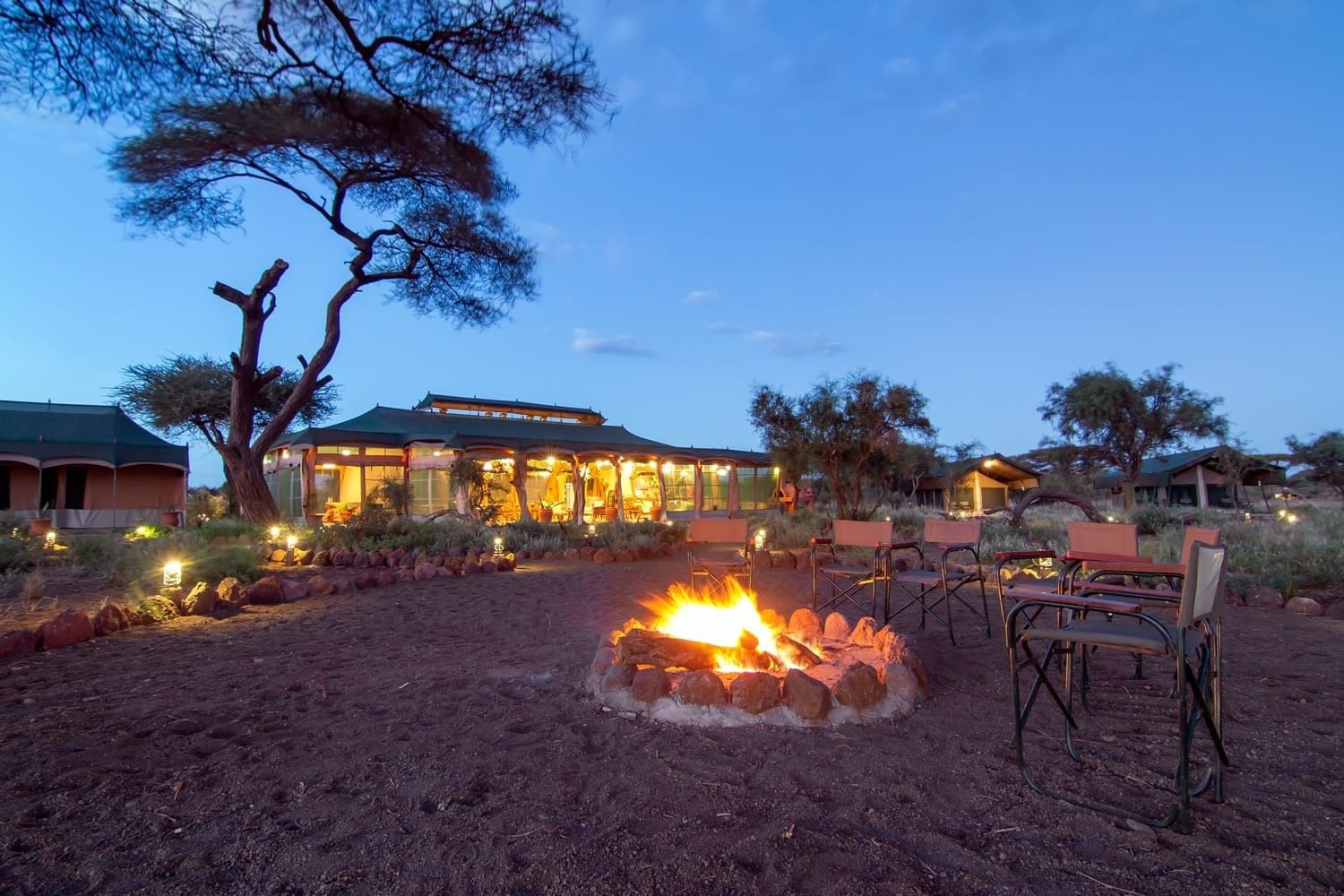Campfire in a stone ring with safari chairs around it in front of the lit main area at Kibo Safari Camp at dusk