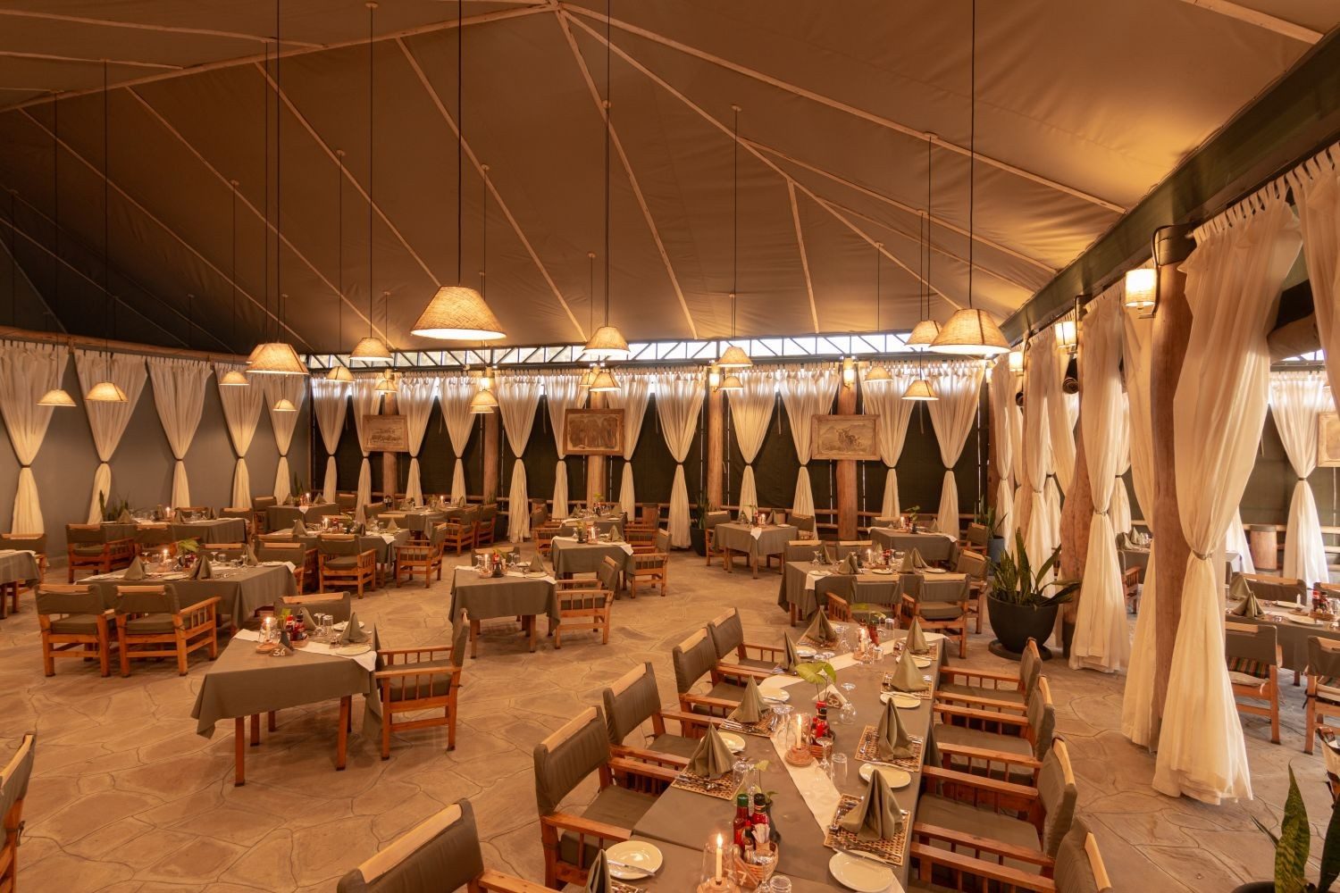 Large dining tent at Kibo Safari Camp with rows of wooden tables and chairs under pendant lights and tied-back curtains