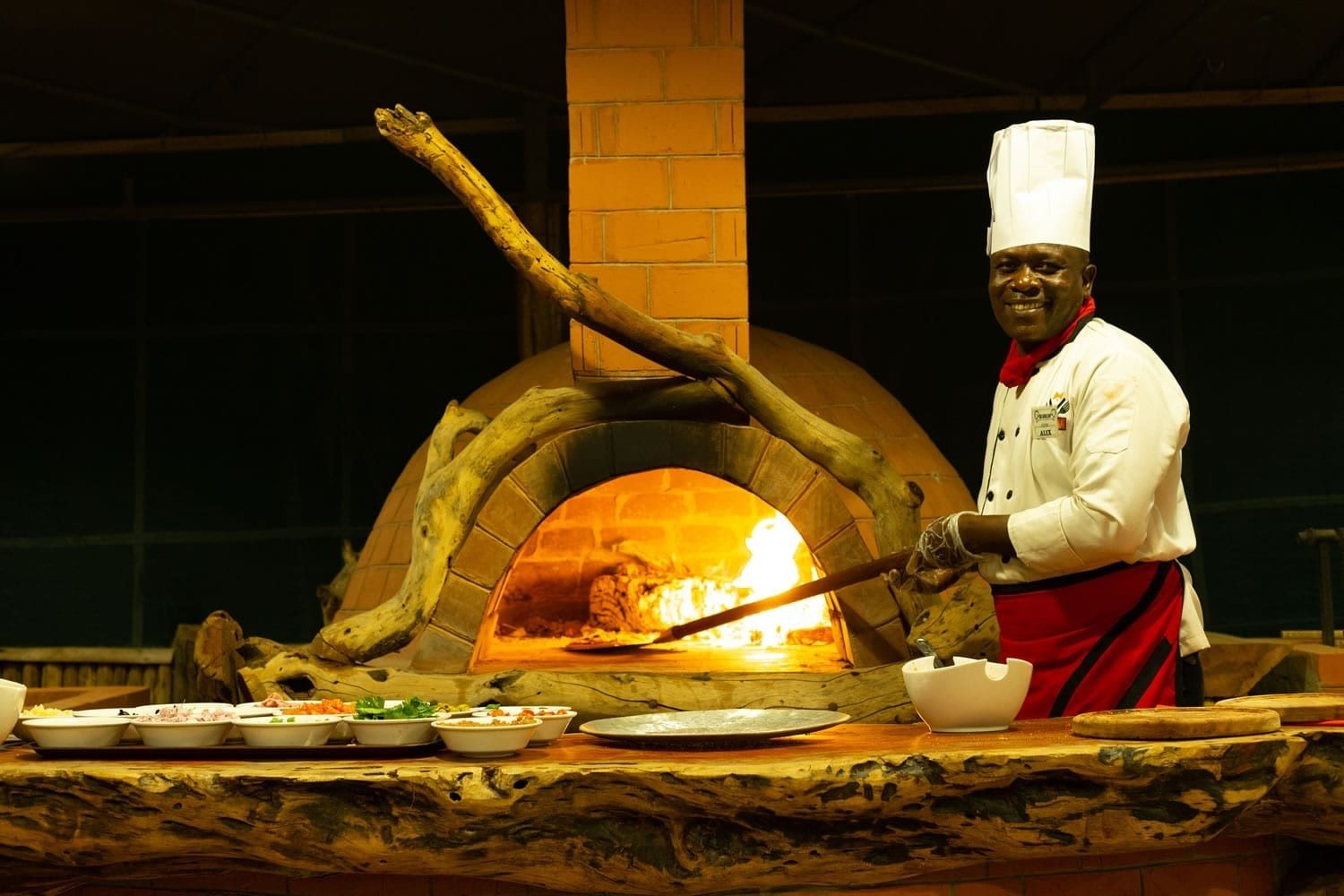 Chef in a white hat sliding a pizza into a wood-fired clay oven at Kibo Safari Camp