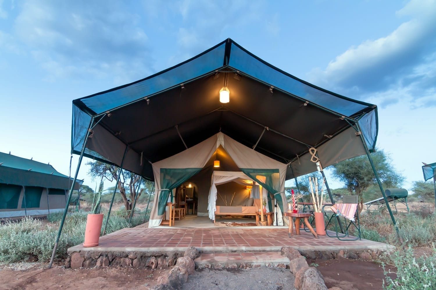 Canvas tent with a lit interior under a high green roof at Kibo Safari Camp, against a blue evening sky