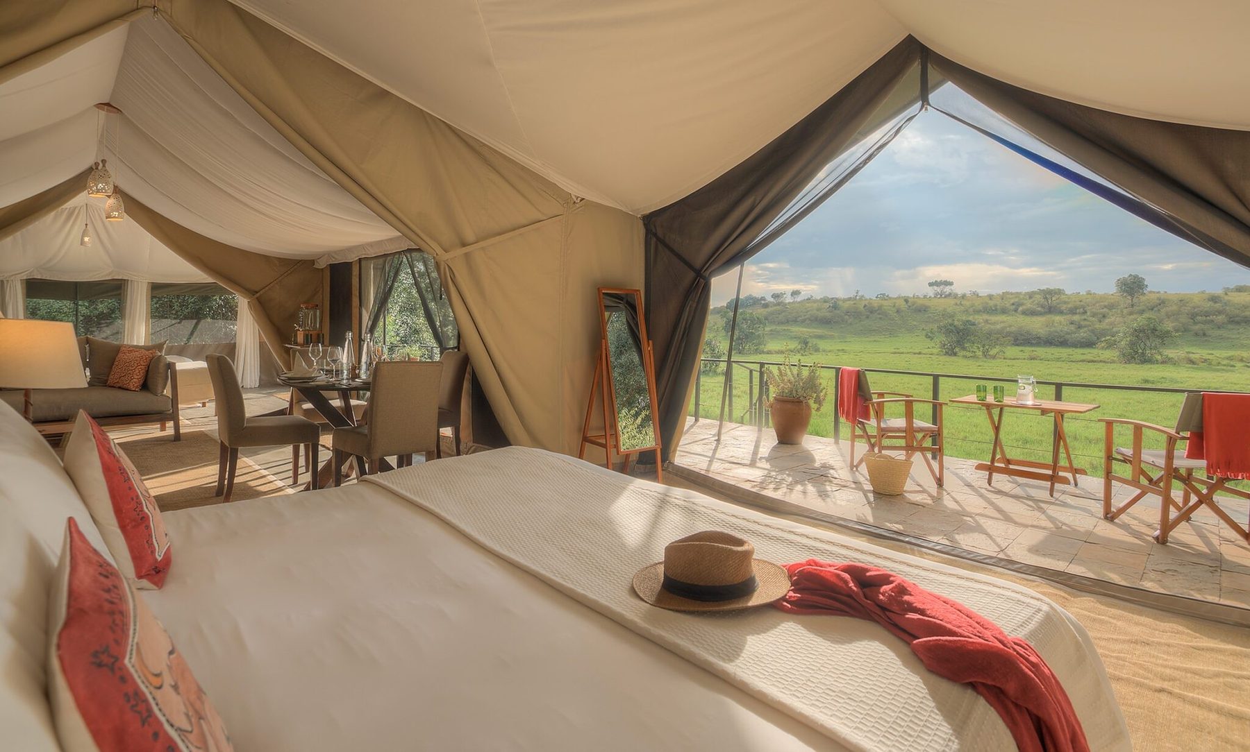Bed with a red throw at Kicheche Mara North, the tent front open to a deck with chairs and green plains