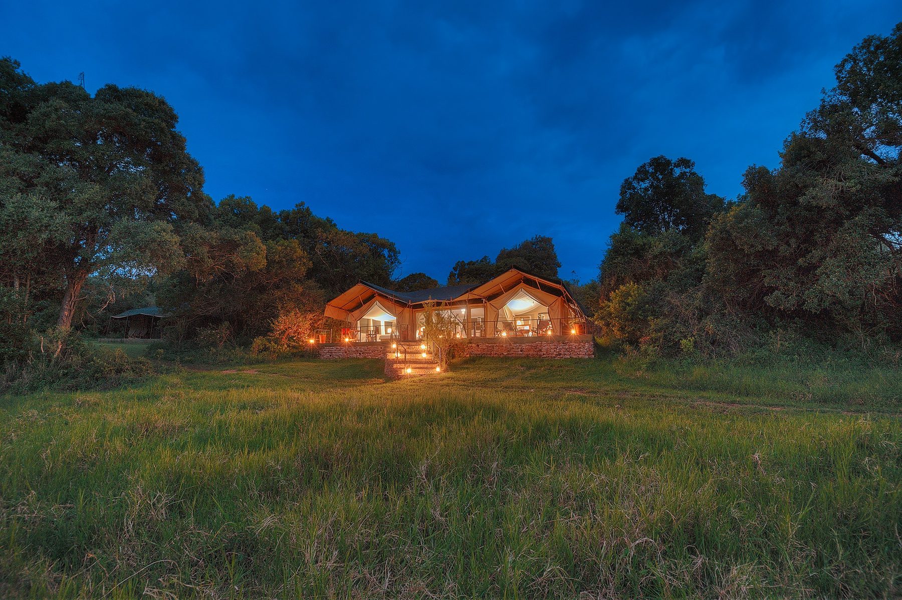 Main tents at Kicheche Mara North lit up at dusk at the end of a wide lawn ringed by trees