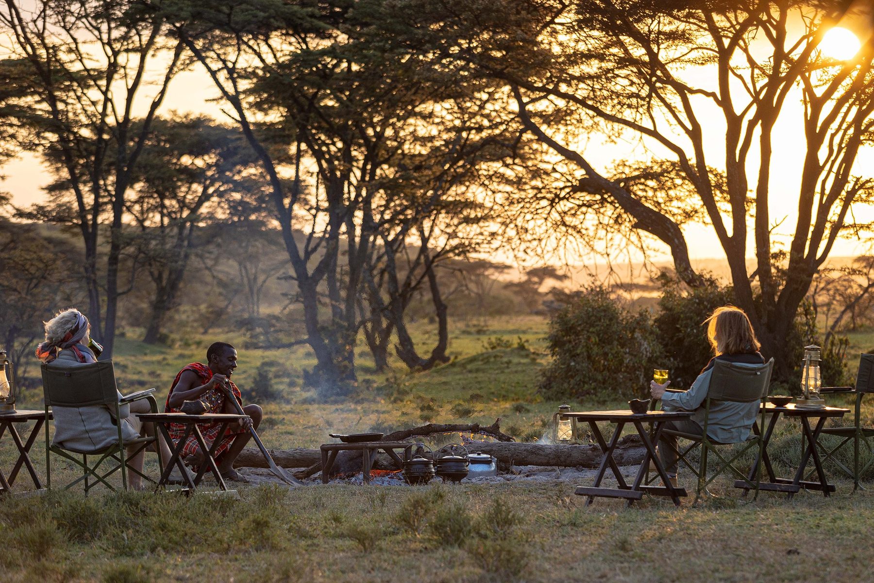 Guests in camp chairs around a fire beneath acacia trees as the sun sets at Kicheche Mara North