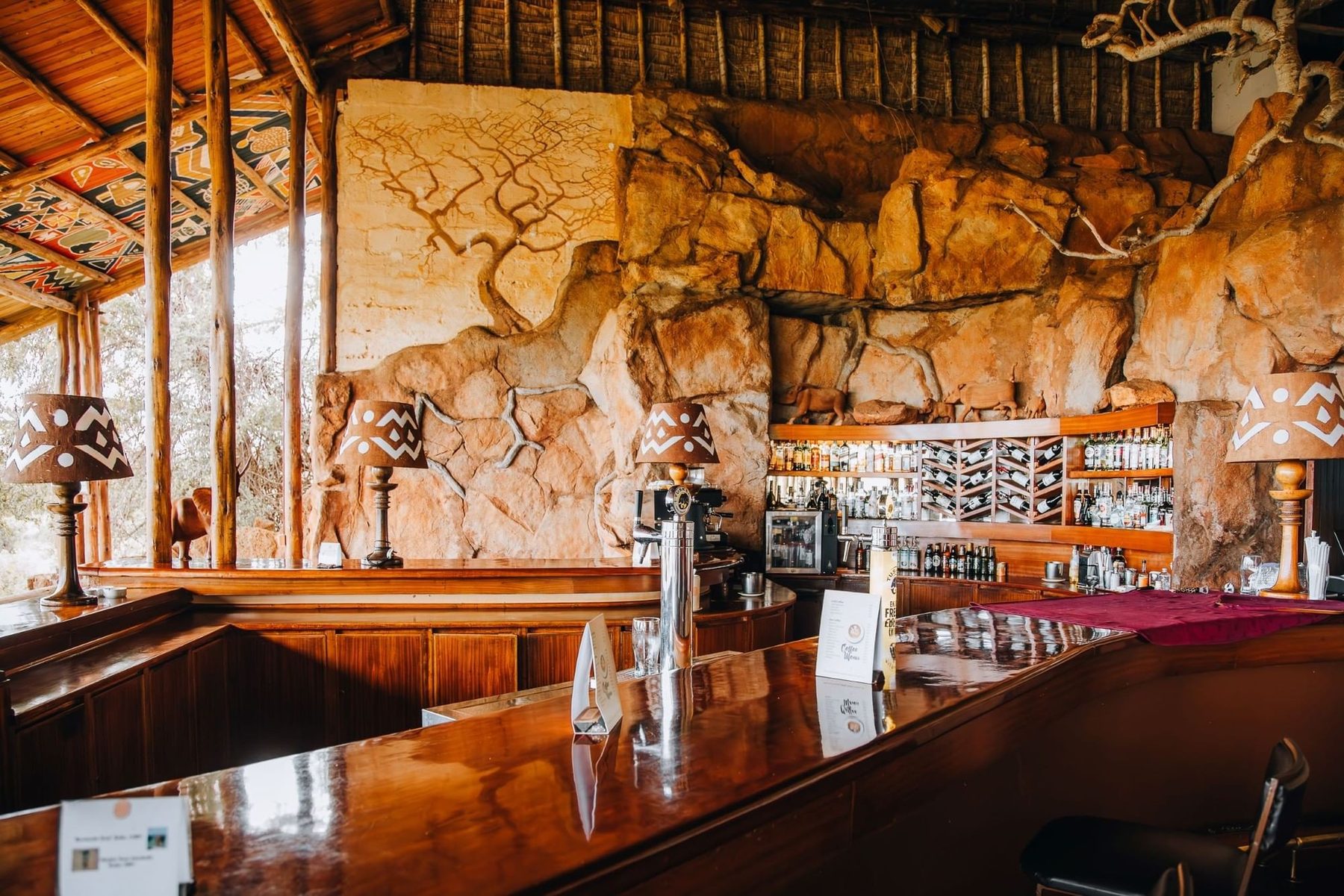 Bar at Kilaguni Serena Safari Lodge with a polished wooden counter and a carved rock wall behind the bottles