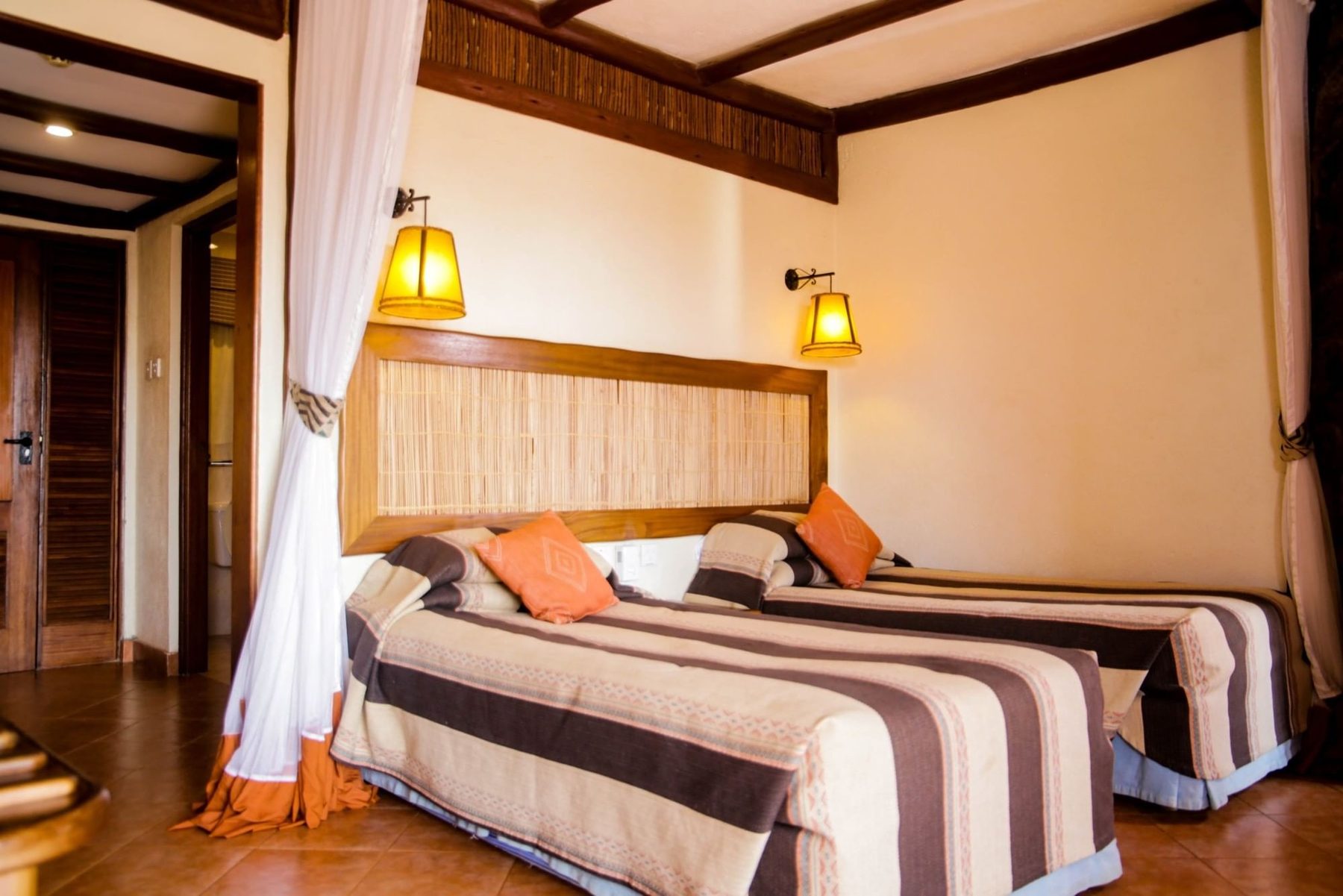 Bedroom at Kilaguni Serena Safari Lodge with a striped bedspread, orange cushions, wall lamps and white curtains