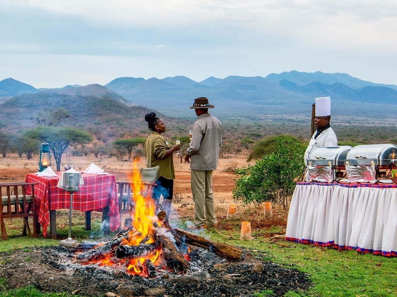 Chef and guests beside a campfire and buffet table set out in the bush, with hills behind in Tsavo West