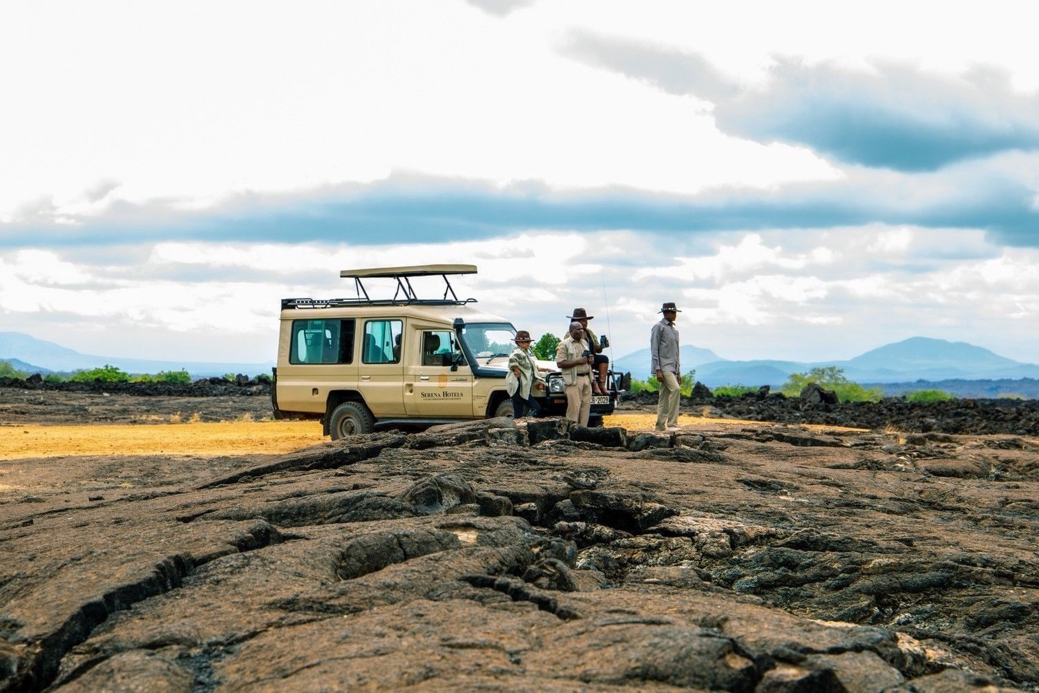 Game drive vehicle parked on black lava rock with guests standing beside it and hills on the horizon in Tsavo West
