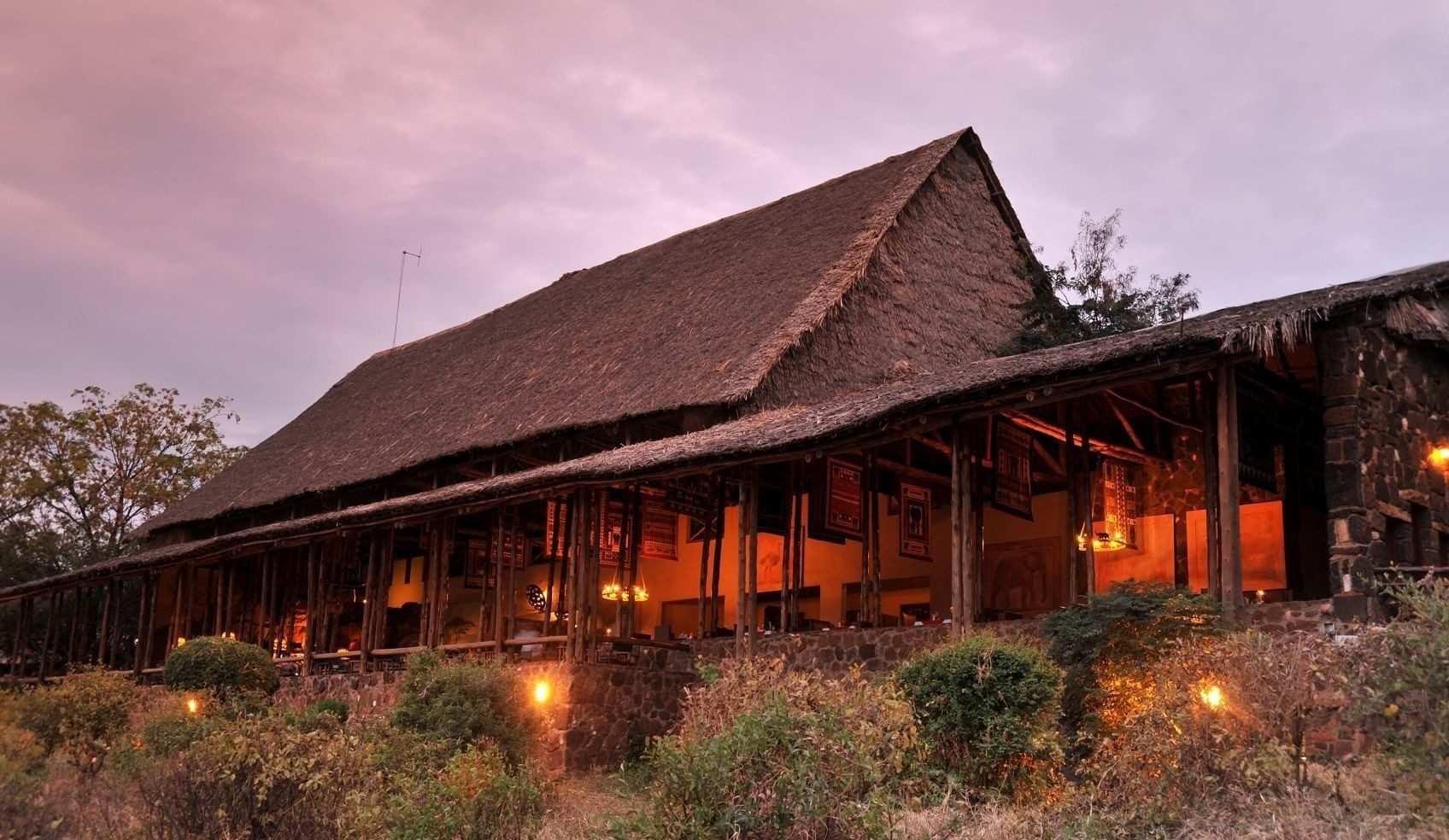 Long thatched main building of Kilaguni Serena Safari Lodge lit up warmly at dusk behind garden plants