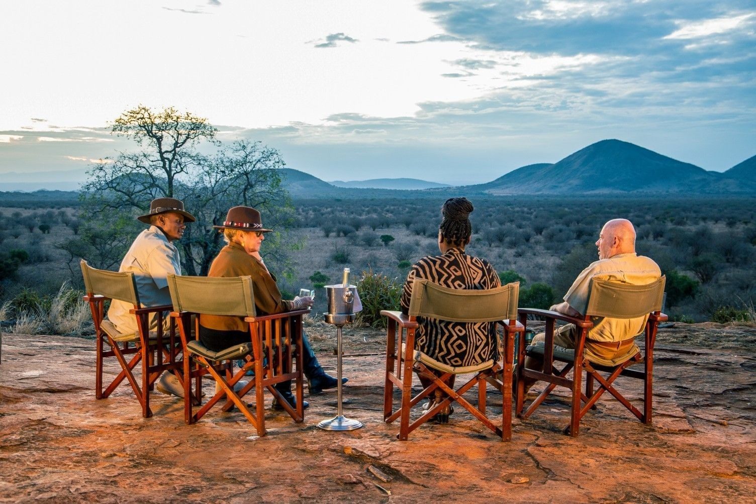 Guests on canvas chairs enjoying sundowners on red earth above the bush, with hills beyond in Tsavo West