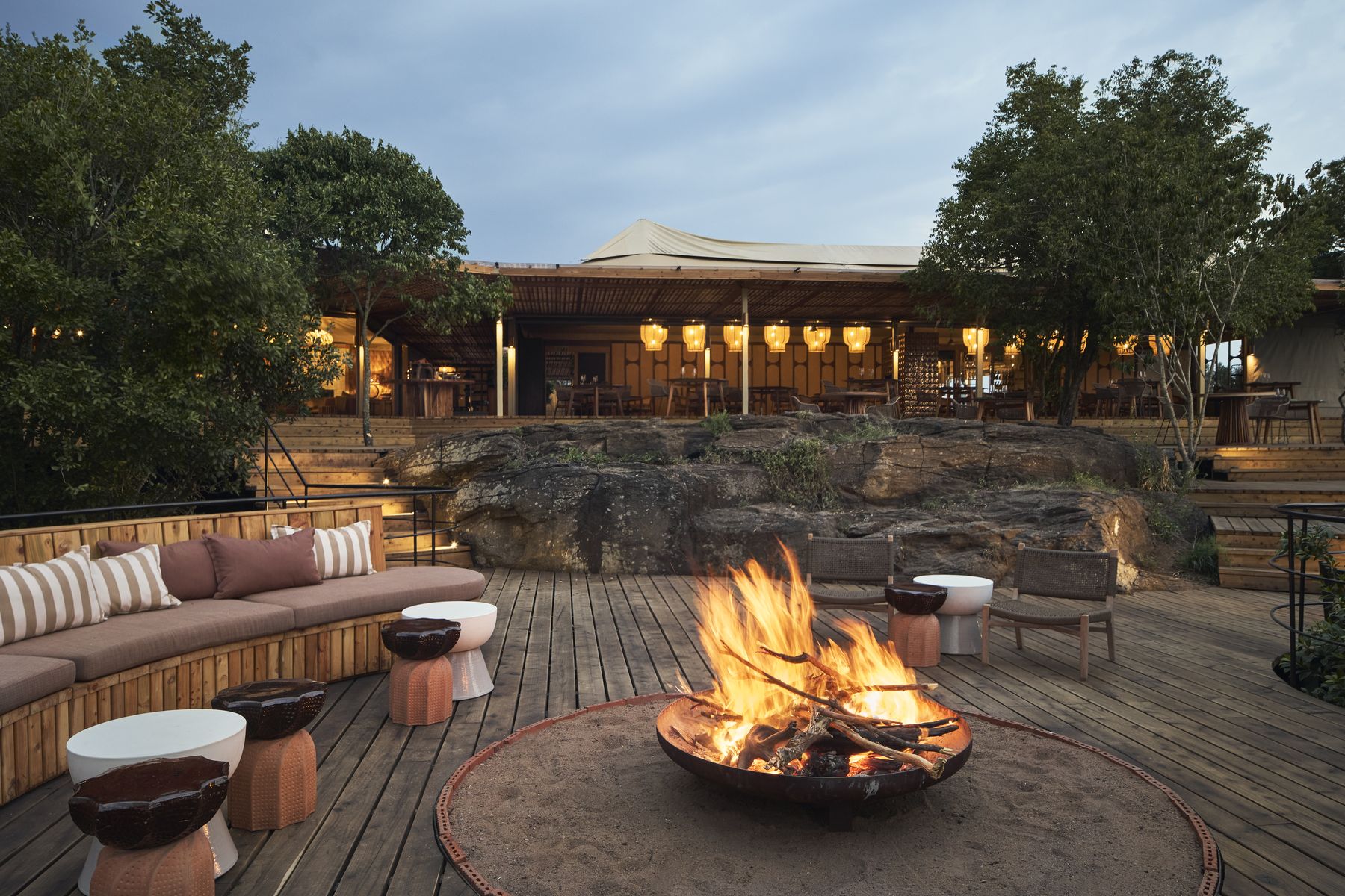 A fire burning in a fire pit on Kinara’s main deck at dusk, with cushioned benches and the lit lounge behind