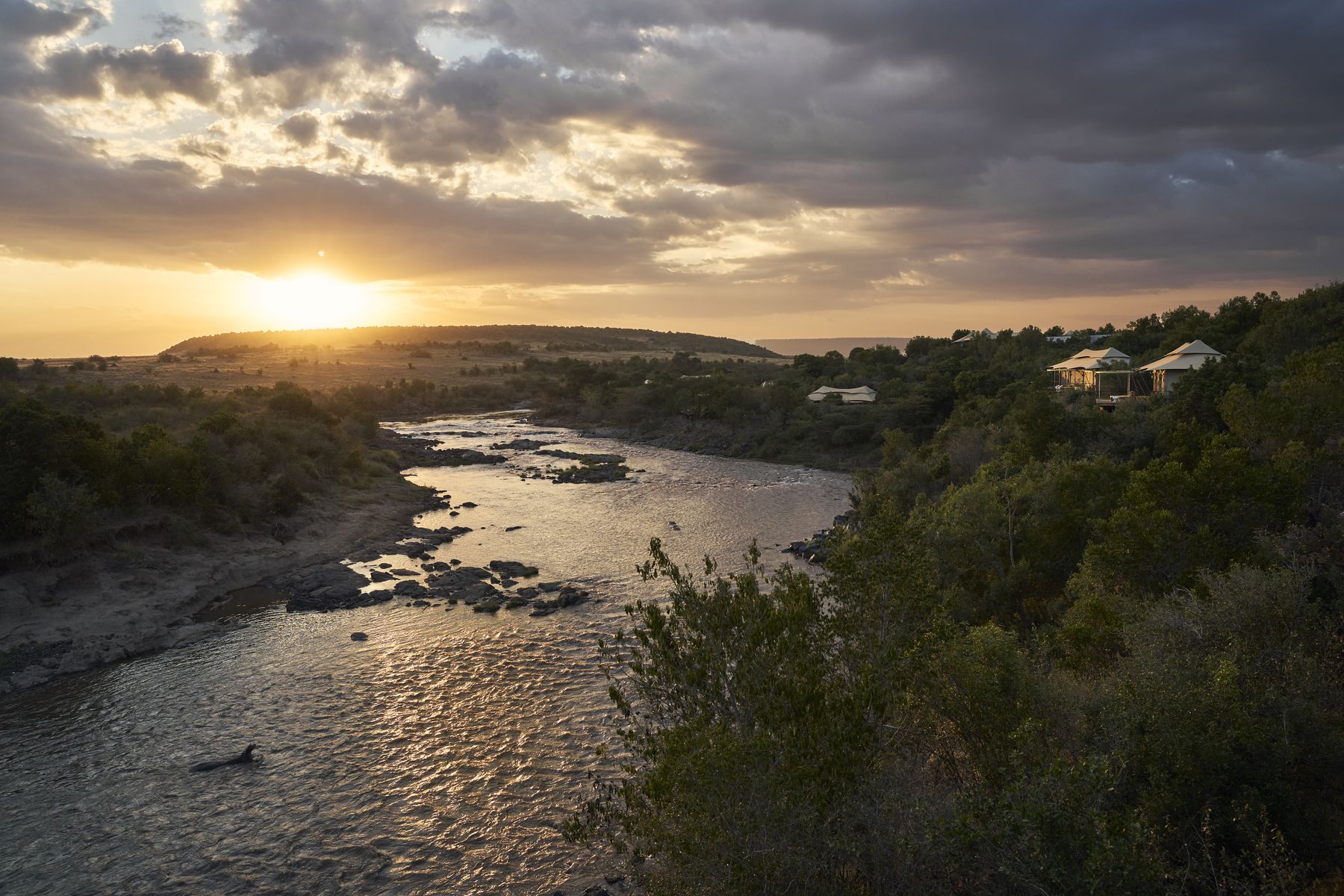 The Mara River flowing over rocks at sunset, with Kinara’s suites on the wooded bank above