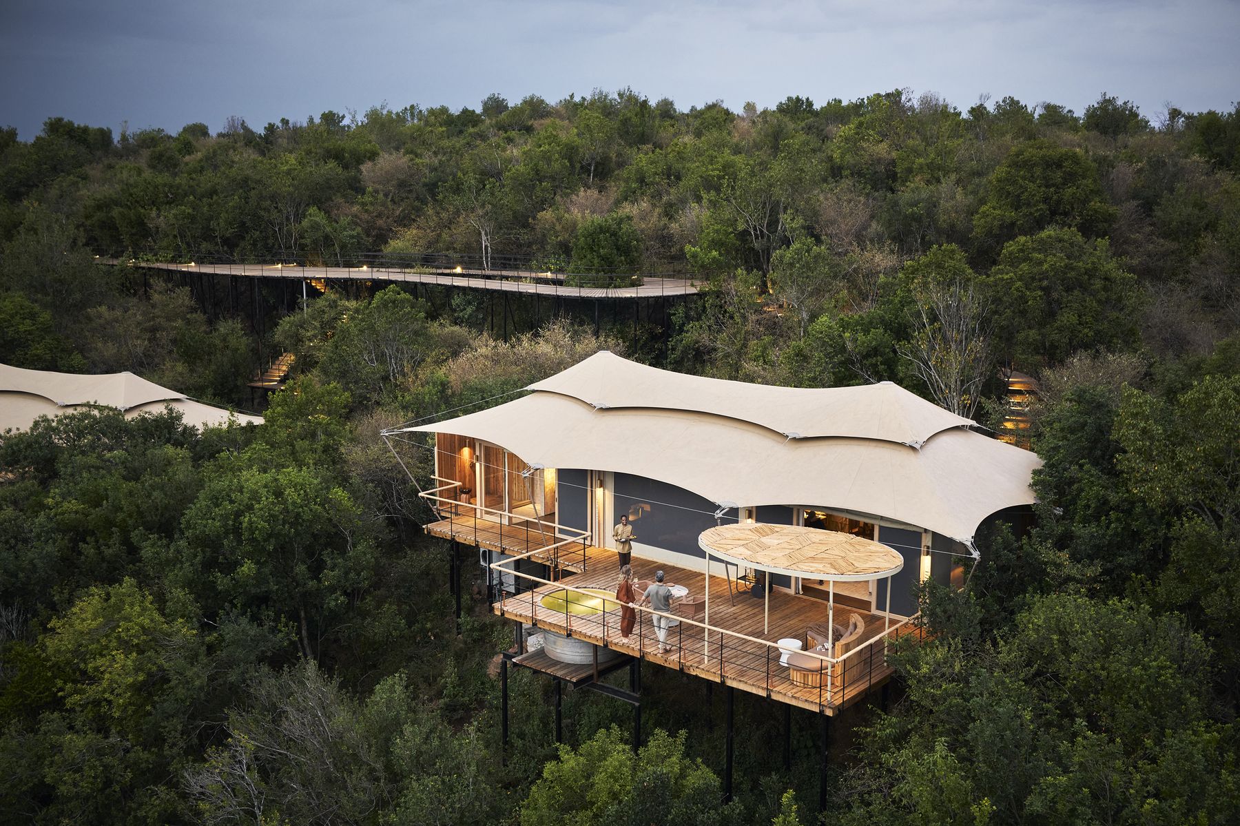 A Kinara suite with a sculpted canvas roof raised among trees, its lit deck seen from above at dusk